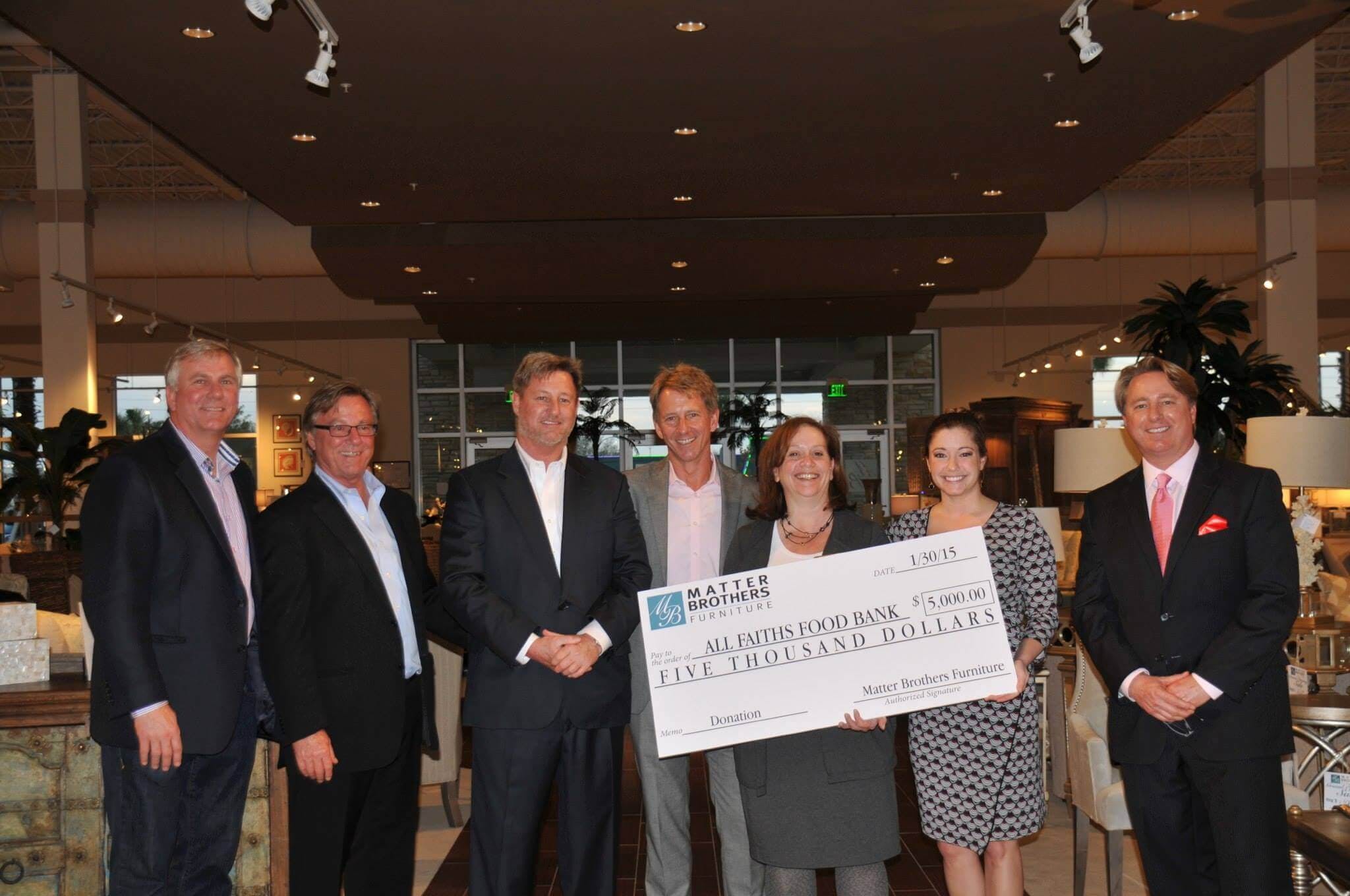 A group of people indoors holding a large check for five thousand dollars from Matter Brothers Furniture to All Faiths Food Bank